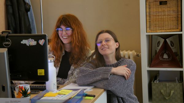 Two people behind a desk, one sitting in a chair and smiling at the computer, and one kneeling with their arms crossed and smiling at the camera