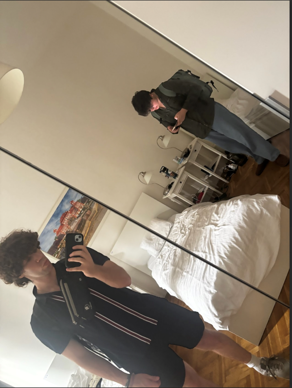 A study abroad student taking a selfie in a mirror with a bed behind them and another study abroad student standing on the other side of the bed looking down at their phone