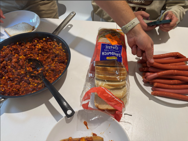 A table full of plates of food including a large dish of chili and a bag of sliced bread, with a hand seen picking up a hot dog from a plate
