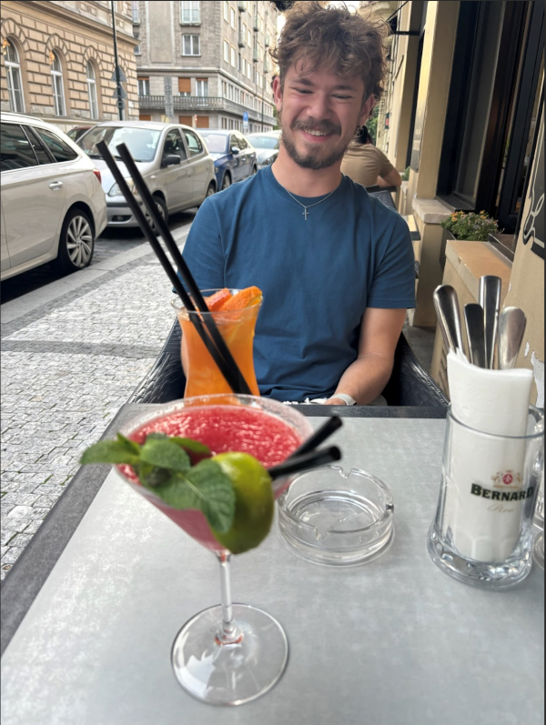 A study abroad student sitting at an outdoor restaurant table and smiling while looking down two brightly-colored drinks on the table