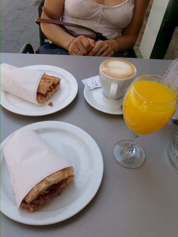 A person seen from the neck down sitting across a small table from the photographer, with plates of sandwiches and a glass of orange juice and a coffee sitting on the table