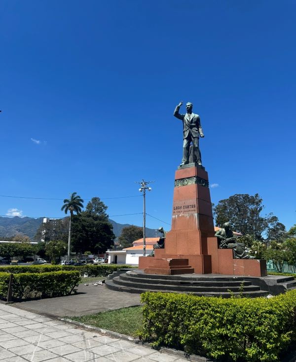 A tall statue of a person in an outdoor greenspace surrounded by several trees and mountains in the distance