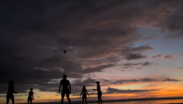 The dark silhouettes of a group of study abroad students playing volleyball on a beach lit by an orange, pink, purple, and blue sunset