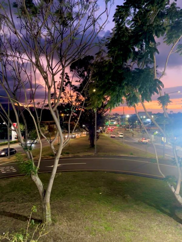 A purple, pink, and orange sunset viewed in the distance behind a grassy green area and several roads with cars driving on them