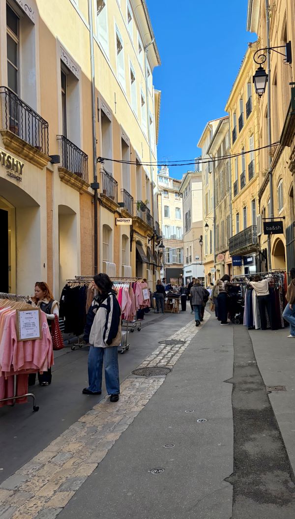 A narrow city street in bright sunlight with people looking at racks of clothes outside shops