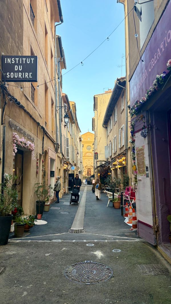A narrow city street in bright sunlight featuring a few people walking about