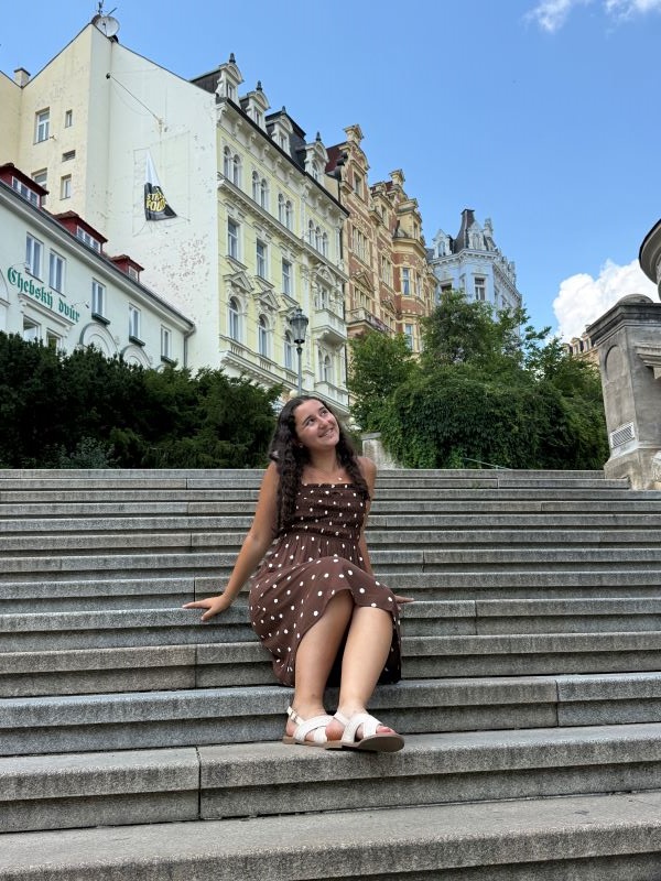 A study abroad student sitting on a stone staircase outdoors and smiling while looking away from the camera