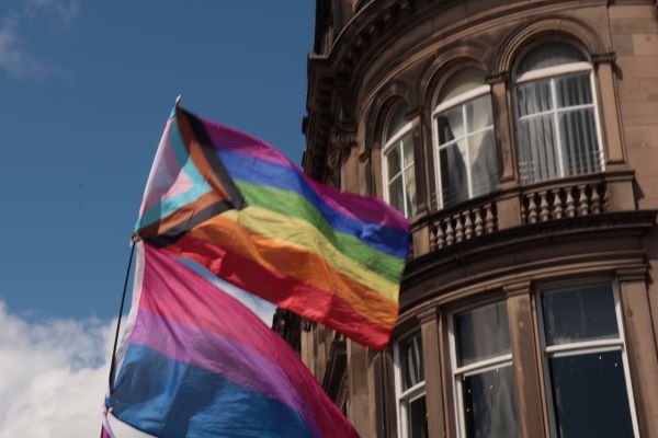 Two brightly-colored Pride flags seen flying in front of a stone building and sunny blue sky