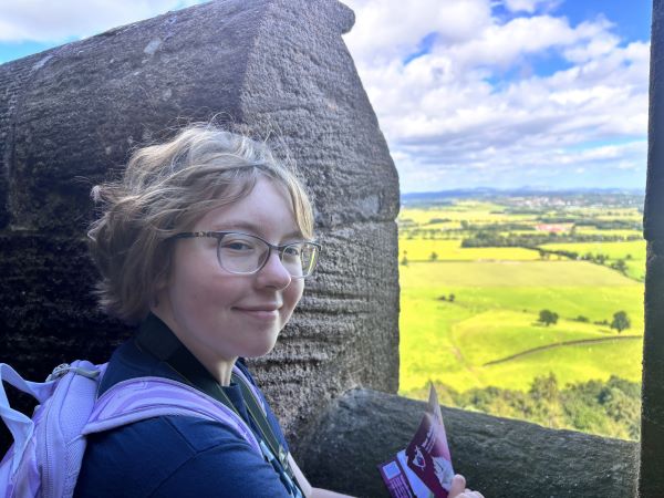 A study abroad student smiling at the camera in front of a stone wall with a bright green landscape in the background