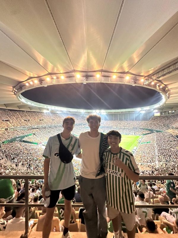 Three study abroad students standing in front of a full soccer stadium and smiling at the camera