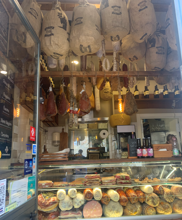 The interior of a small sandwhich shop, featuring a glass counter with many types of meat and sandwiches