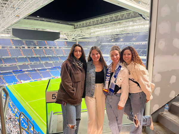 four students smile for picture in a soccer stadium with the field behind them.