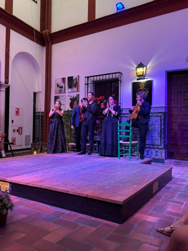 A group of flamenco performers dressed in traditional black outfits, clapping their hands in front of a small stage