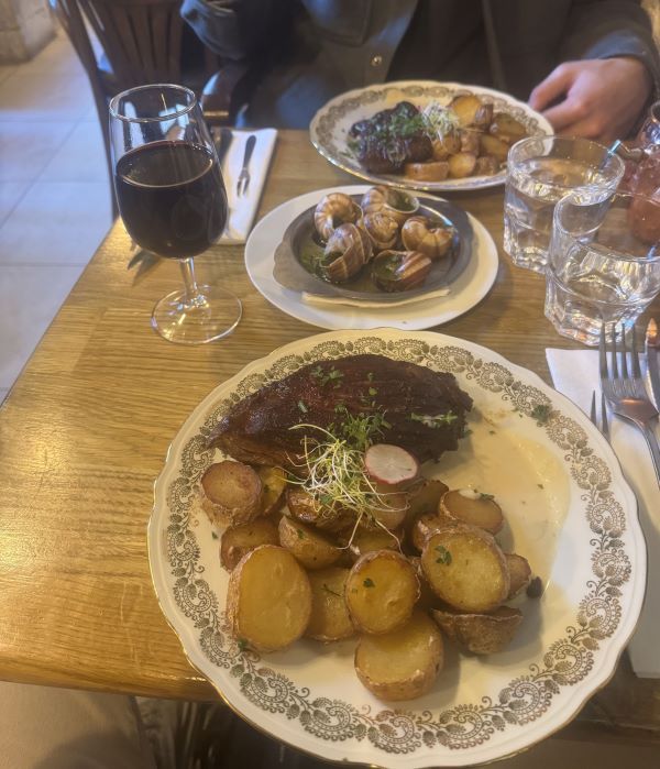 A piece of cooked meat and small round potatoes sitting on an ornate white plate on a wooden restaurant tabletop