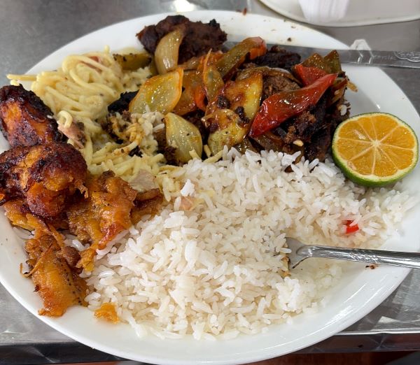 A white plate full of food sitting on a table, featuring rice, meat, cooked vegetables, and half of a citrus fruit