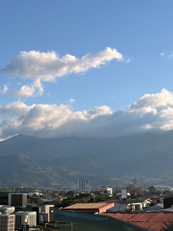 A large green mountain seen in the distance under fluffy white clouds and a blue sky, with city buildings in the near distance