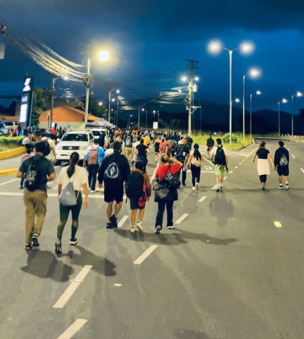 A group of people as seen from behind walking on a brightly-lit road toward mountains in the distance at night