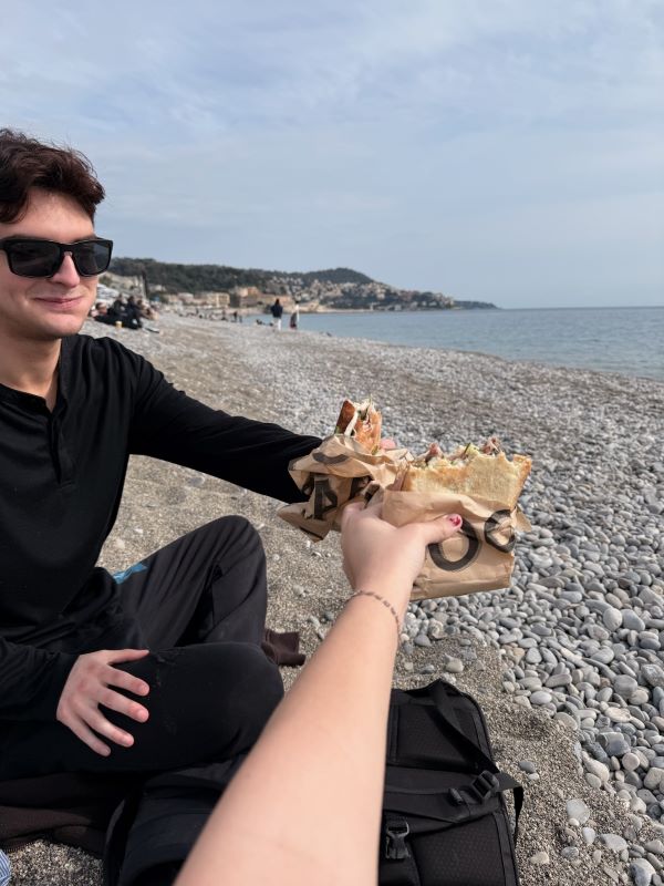 An arm holding out a sandwich to meet the outheld sandwich of a study abroad student wearing black clothes and sunglasses sitting on a rocky beach shore