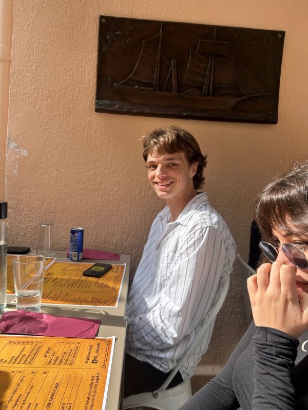 A study abroad student bathed in sunlight and sitting at an outdoor restaurant table and turned to the side to smile at the camera