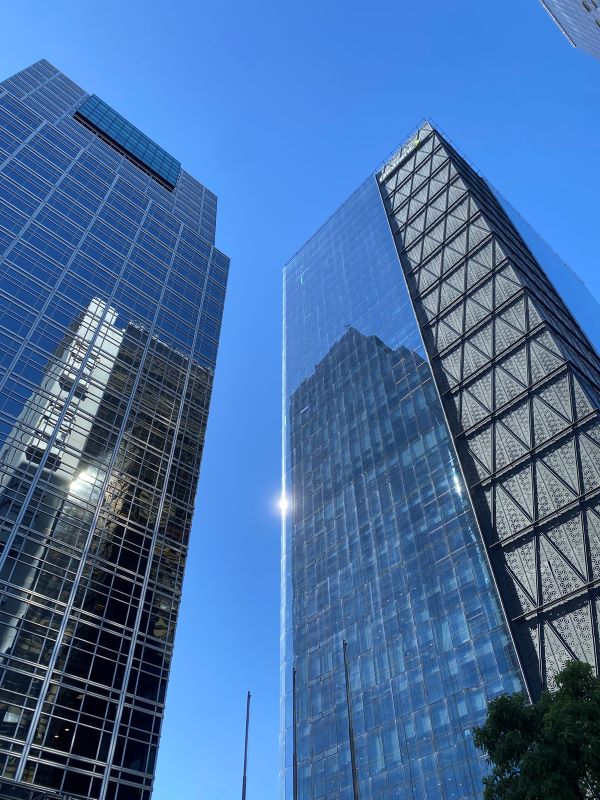 Two tall modern glass-and-steel buildings as seen from below against a bright blue sky
