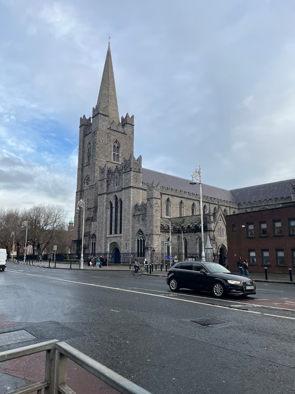 A large gray stone cathedral sitting behind a road with cars driving on it underneath a cloudy blue sky
