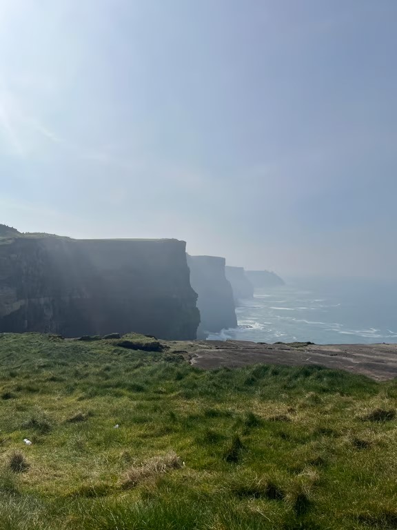 A large cliff formation seen on the left side next to a large body of water with a grassy green slope in the foreground