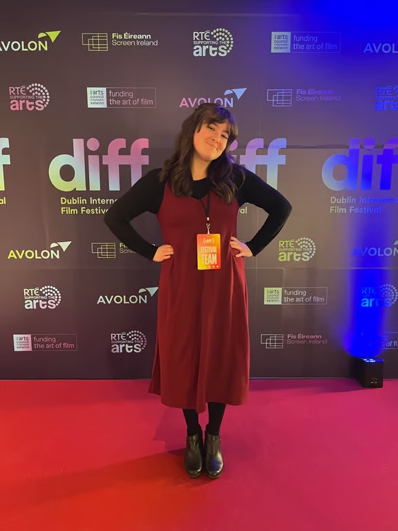 A study abroad student smiling and looking at the camera with their hands on their hips and wearing a red dress standing in front of a wall with many logos on it