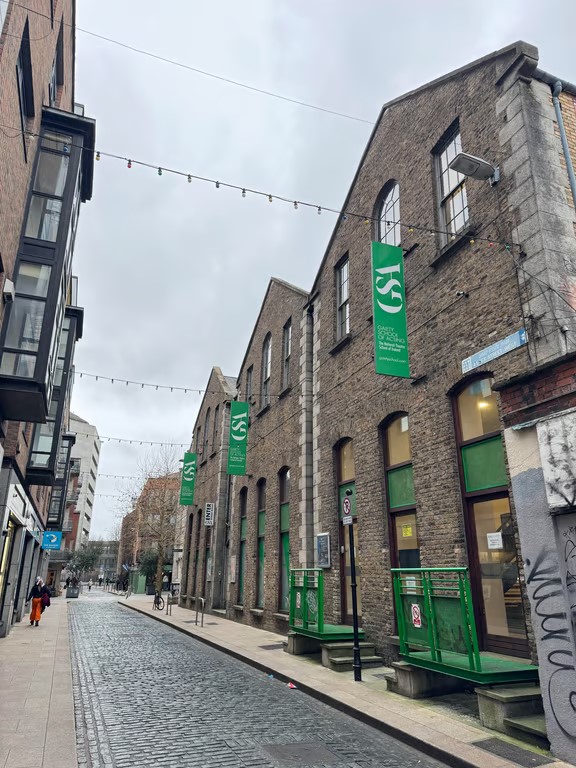 A cobblestone city street with tall stone buildings to the right featuring green flags reading