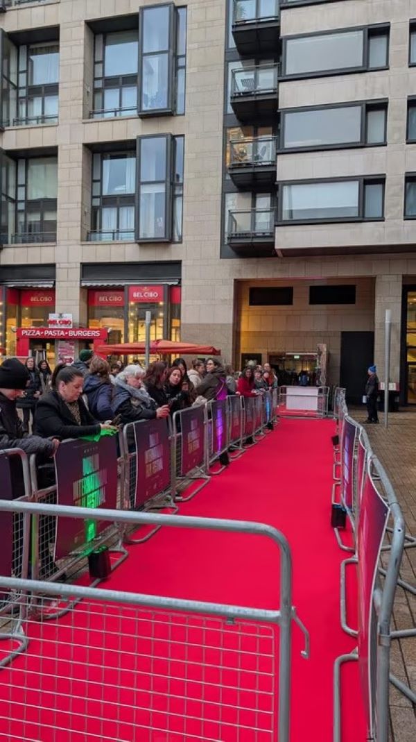 A red carpet leading into a tall building, with people standing to the left of it behind a short barrier.