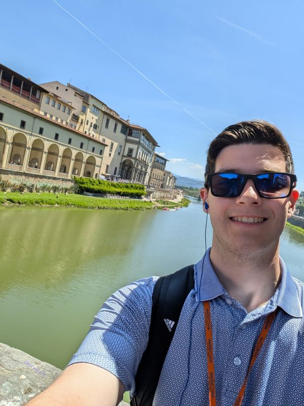 A study abroad student wearing a blue polo shirt and sunglasses smiling and taking a selfie in front of a river hemmed in by city buildings