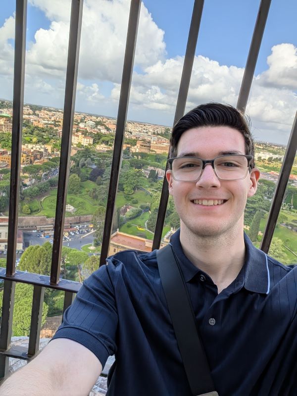 A study abroad student wearing glasses and a dark polo shirt smiling and taking a selfie in front of a city view seen through thin black bars