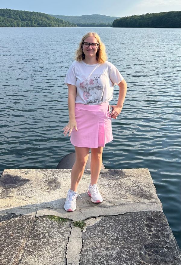 A study abroad student with blonde hair and glasses standing on a rocky structure outdoors in front of a large body of water and smiling at the camera