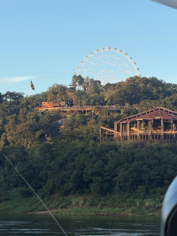 A green tree-filled hill seen across a body of water and featuring several buildings and a Ferris wheel in the background
