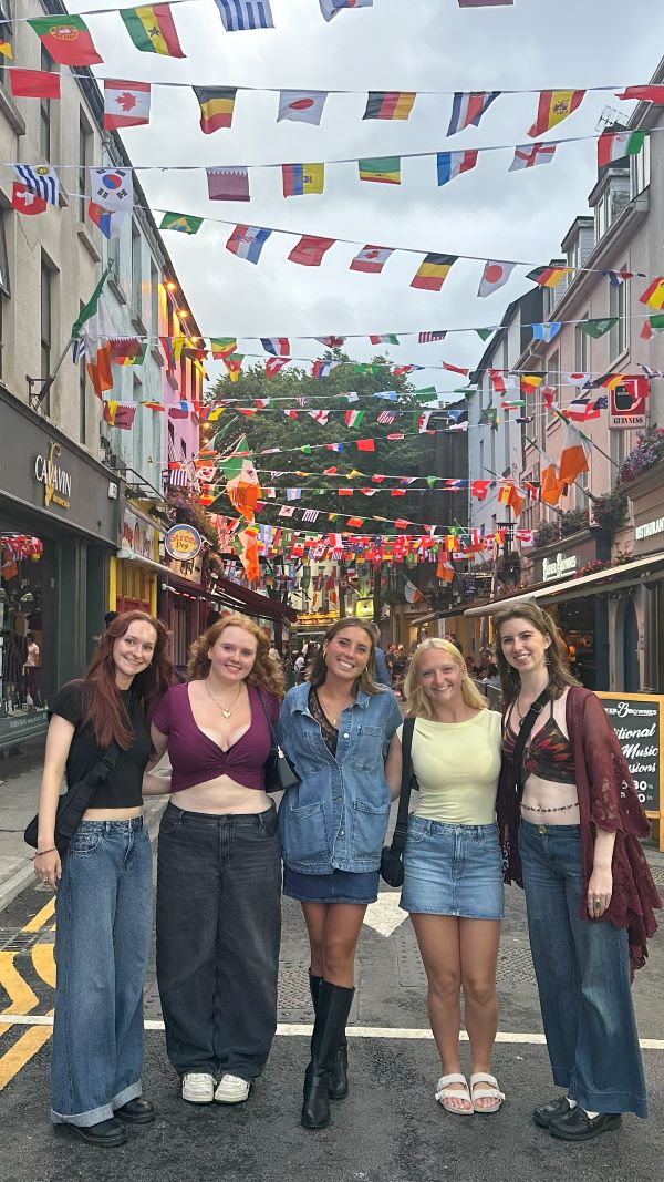 Five study abroad students standing outdoors on a city street underneath colorful banners strung between two buildings overhead and smiling at the camera