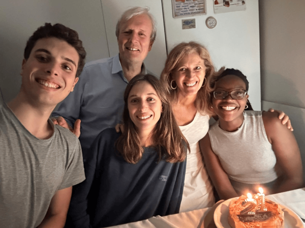 Five people taking a selfie in an indoor setting while on the table in front of them sits a cake with