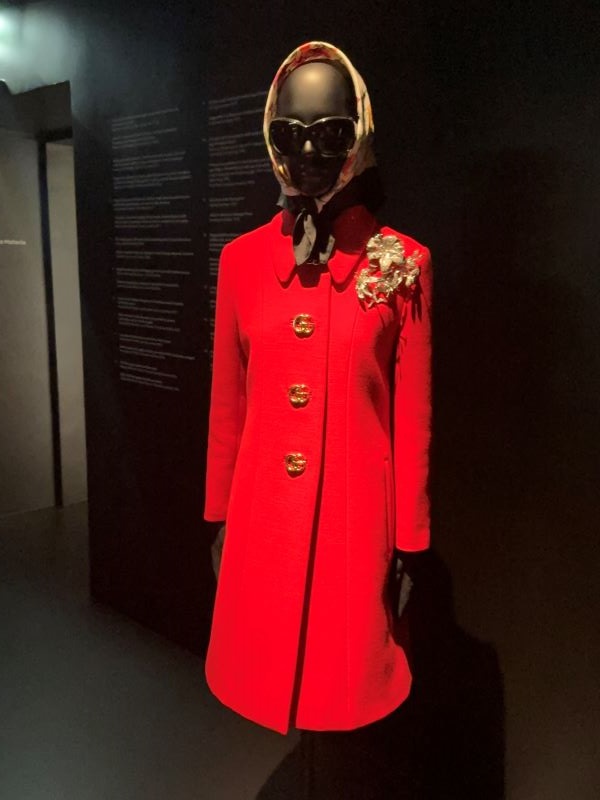 A mannequin seen indoors draped in a bright orange-red coat and wearing a headscarf and sunglasses