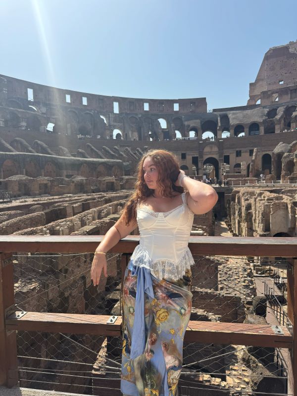 A study abroad student wearing a white top and brightly colored skirt standing in front of a large stone structure and posing while looking away from the camera