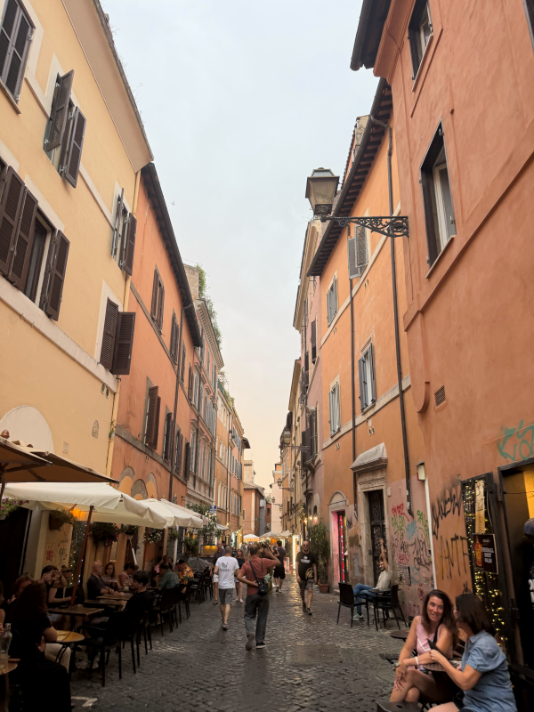 People walking along and sitting around on a city street hemmed in by tall sand-colored buildings