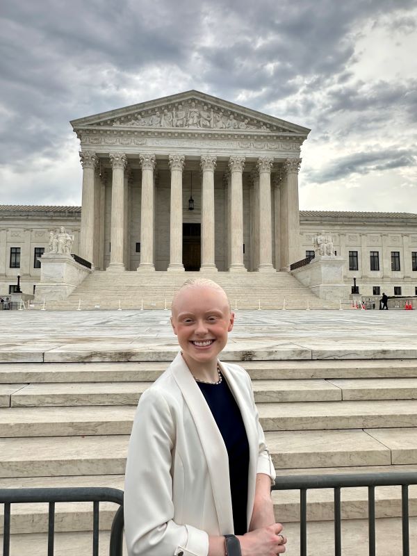 A pre-law student wearing a black top and white blazer standing in front of a large ornate building and smiling at the camera