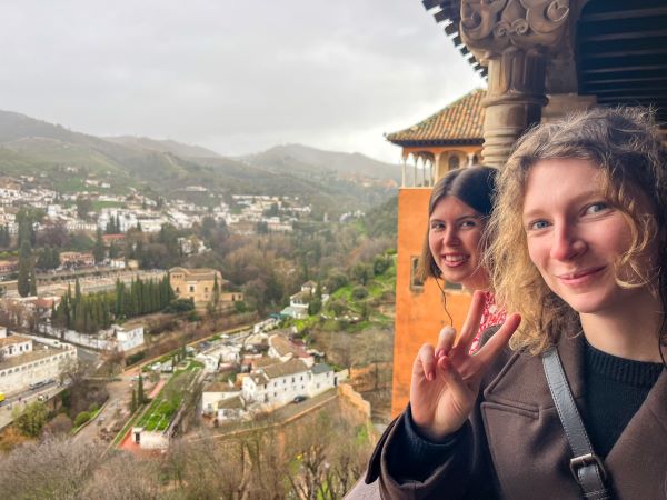 Two study abroad students smiling and taking a selfie in front of a view of city buildings