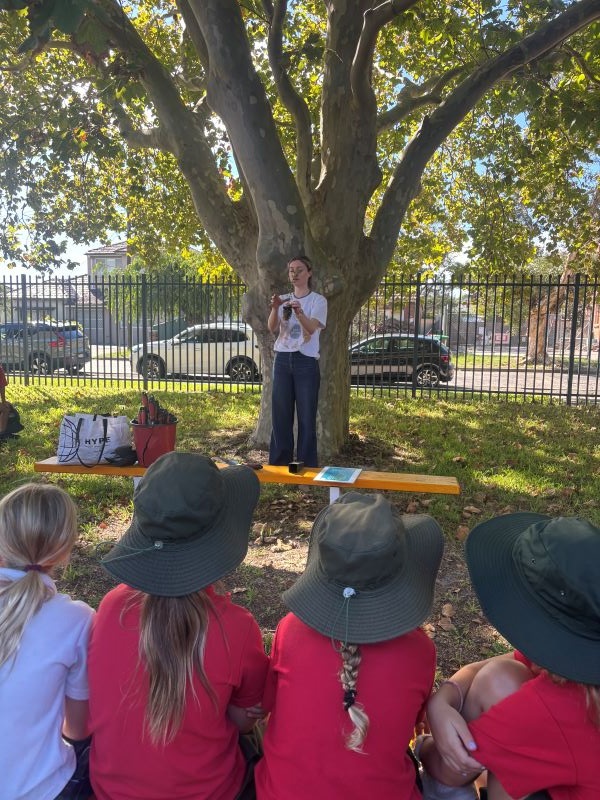 A person standing outdoors and speaking to several children who are only seen from behind