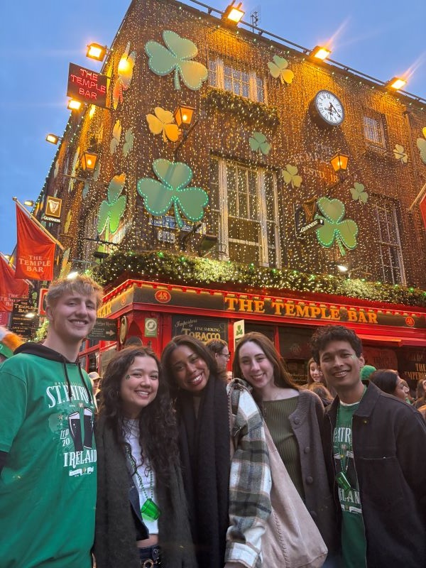 Five study abroad students dressed in green and standing outdoors smiling at the camera in front of a building with the words
