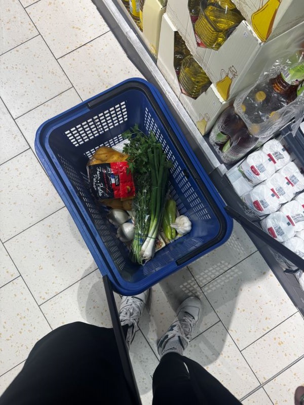 A pair of legs in black trousers standing next to a small blue grocery store basket seen from above and filled with produce