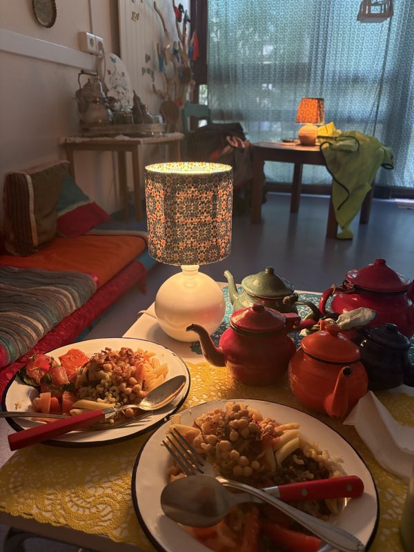 A table in an indoor setting full of plates of food, several colorful teapots, and a lamp