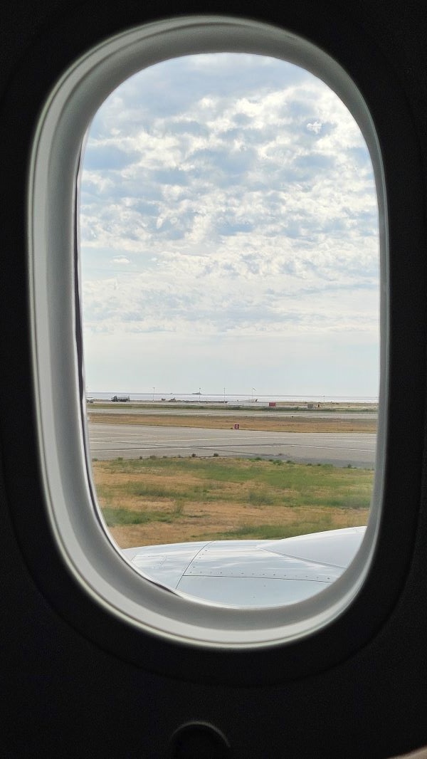A view through an oval-shaped airplane window of an airport landing strip and a cloudy blue sky
