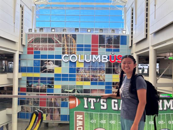 A study abroad student wearing a backpack and smiling at the camera in front of a sign reading