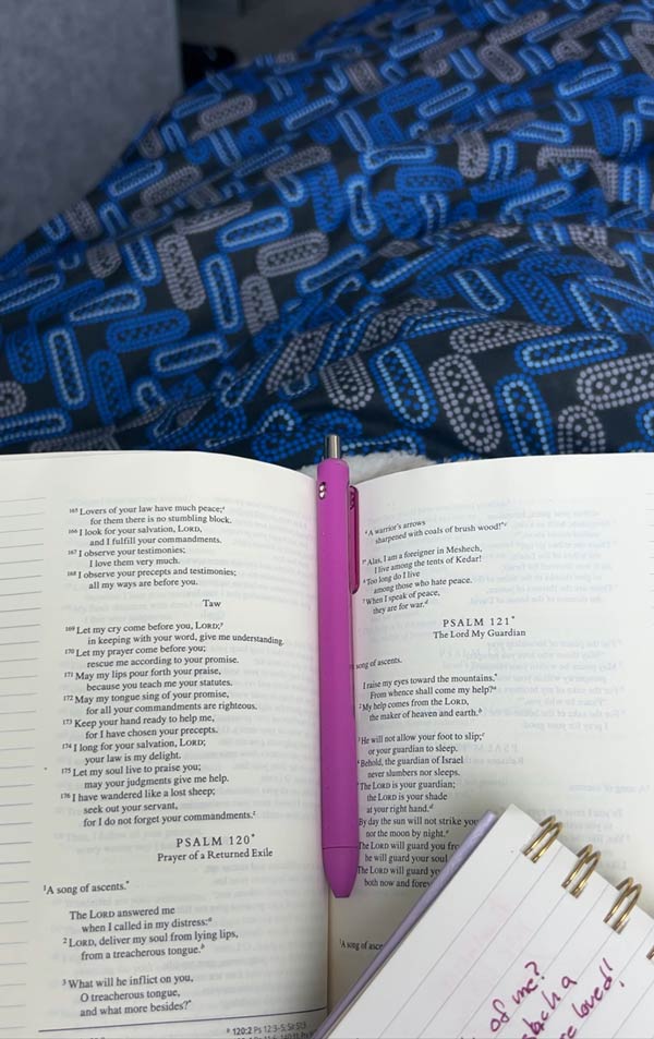 A Bible laid open to two pages with a notebook page in the corner and a pen in the middle of the book