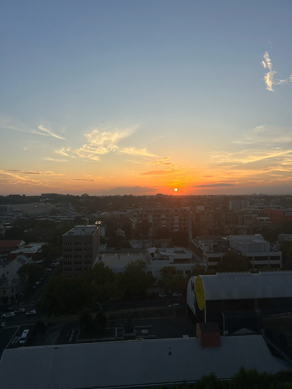 A view from a window of buildings underneath an orange sun setting in the sky