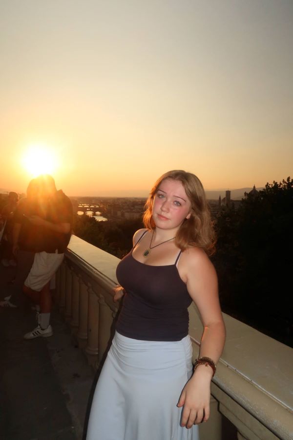 A study abroad student with shoulder-length blonde hair leaning against a stone wall during sunset and looking at the camera
