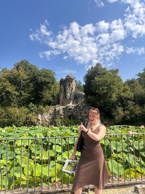 A study abroad student wearing a brown dress standing in front of a small fence and pointing to a large statue of a man in the background while smiling at the camera